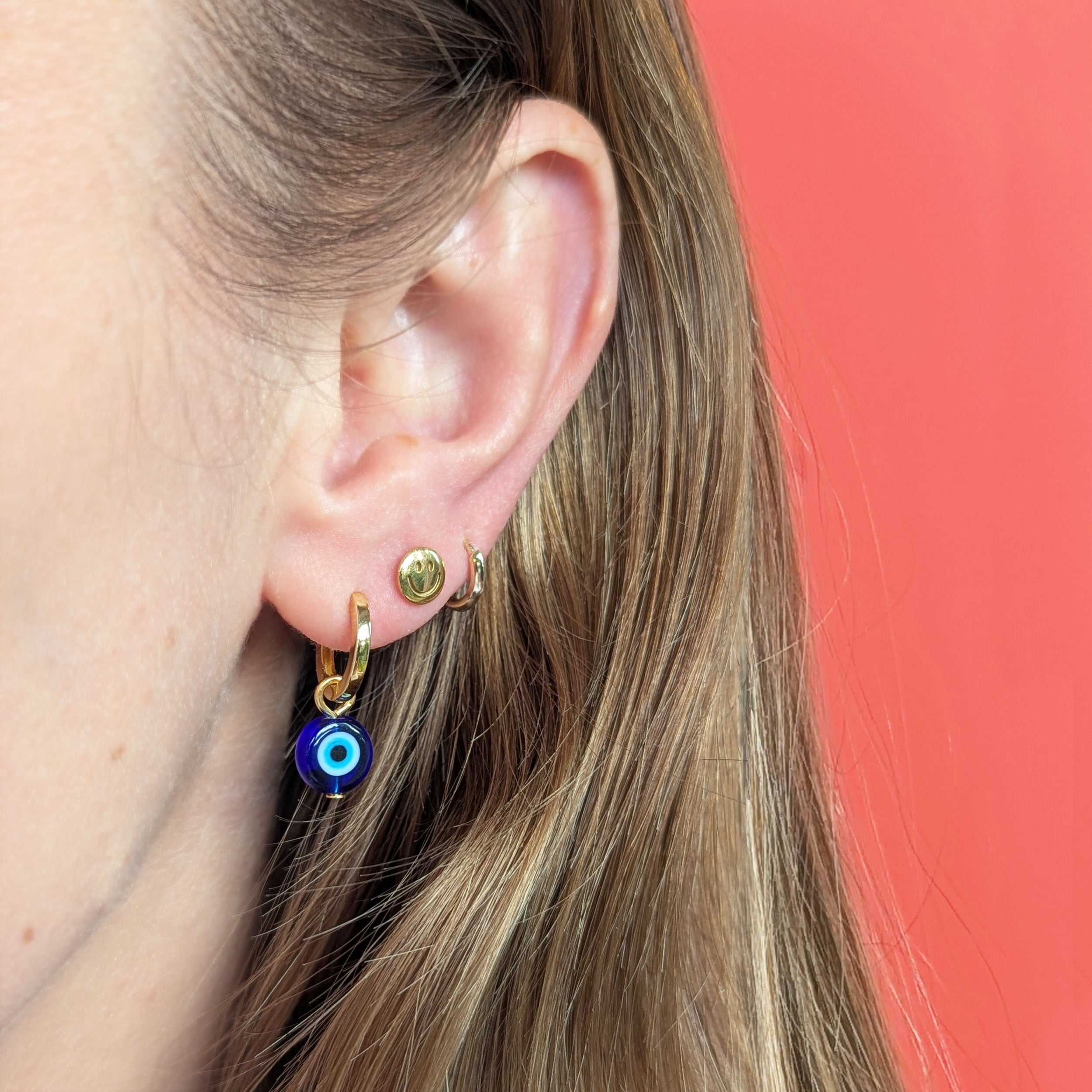 Gold hoop earring with blue evil eye charm worn by a person with a coral background.