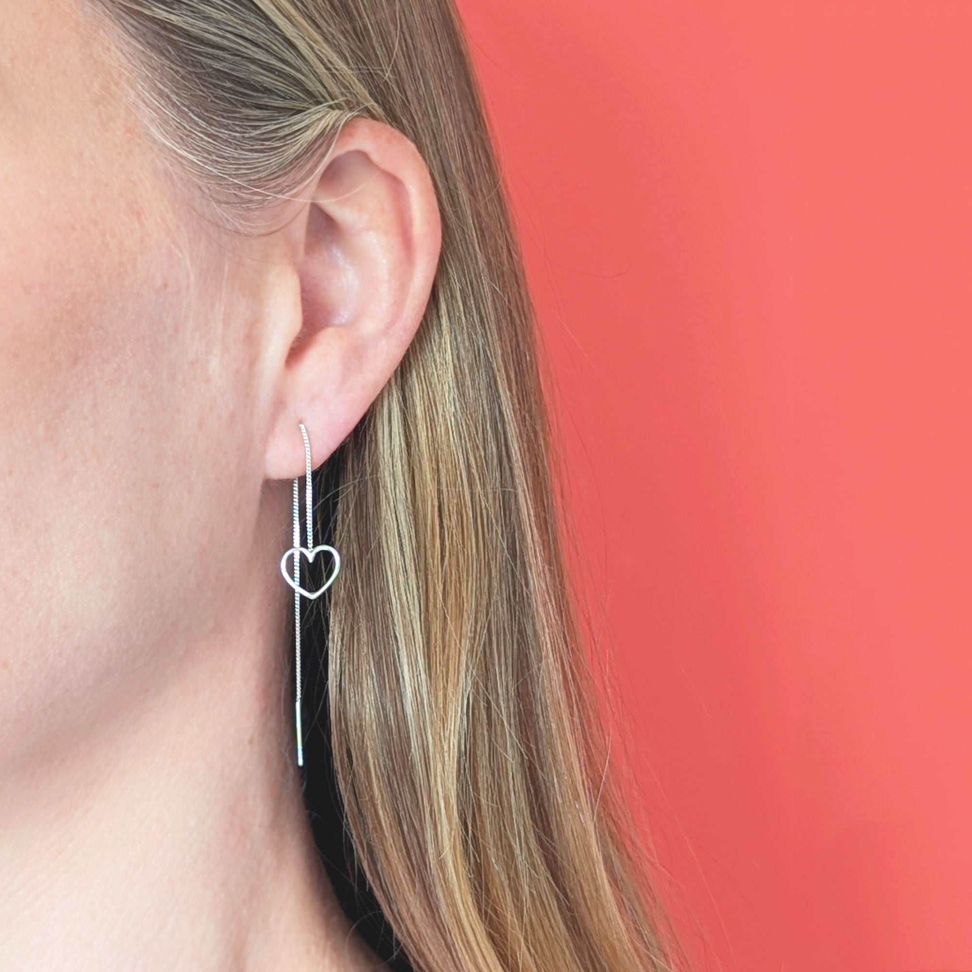Silver heart-shaped chain threader earring worn by a person with a coral background