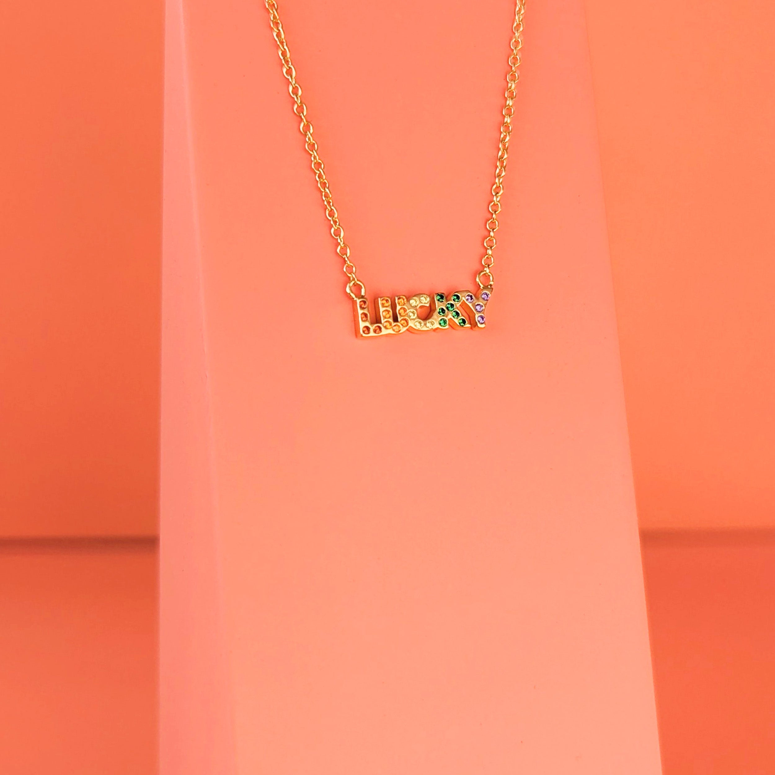 Gold necklace with 'LUCKY' set with rainbow gemstones on a coral background