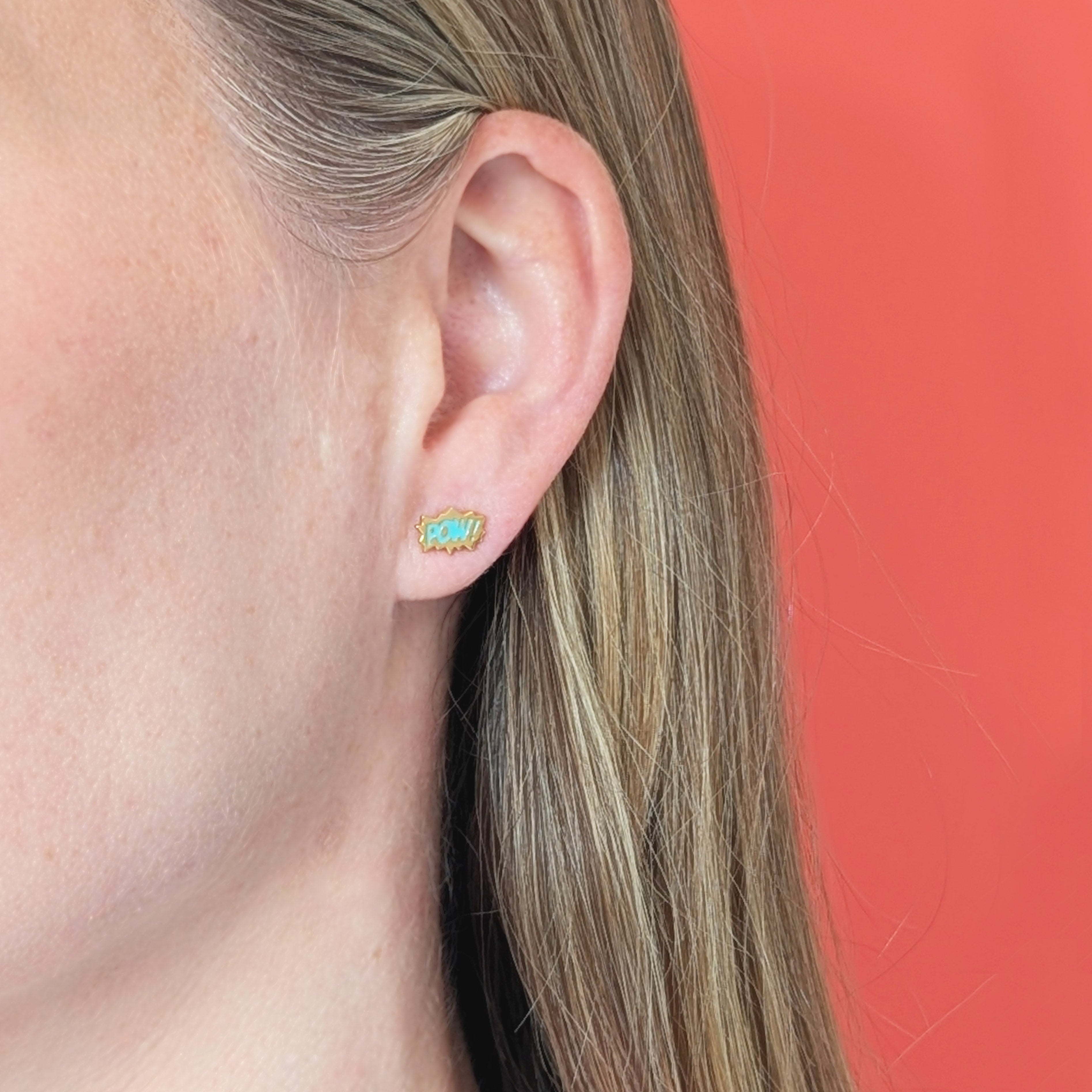 Close-up of an ear wearing a gold comic burst earring with turquoise BAM! and coral background