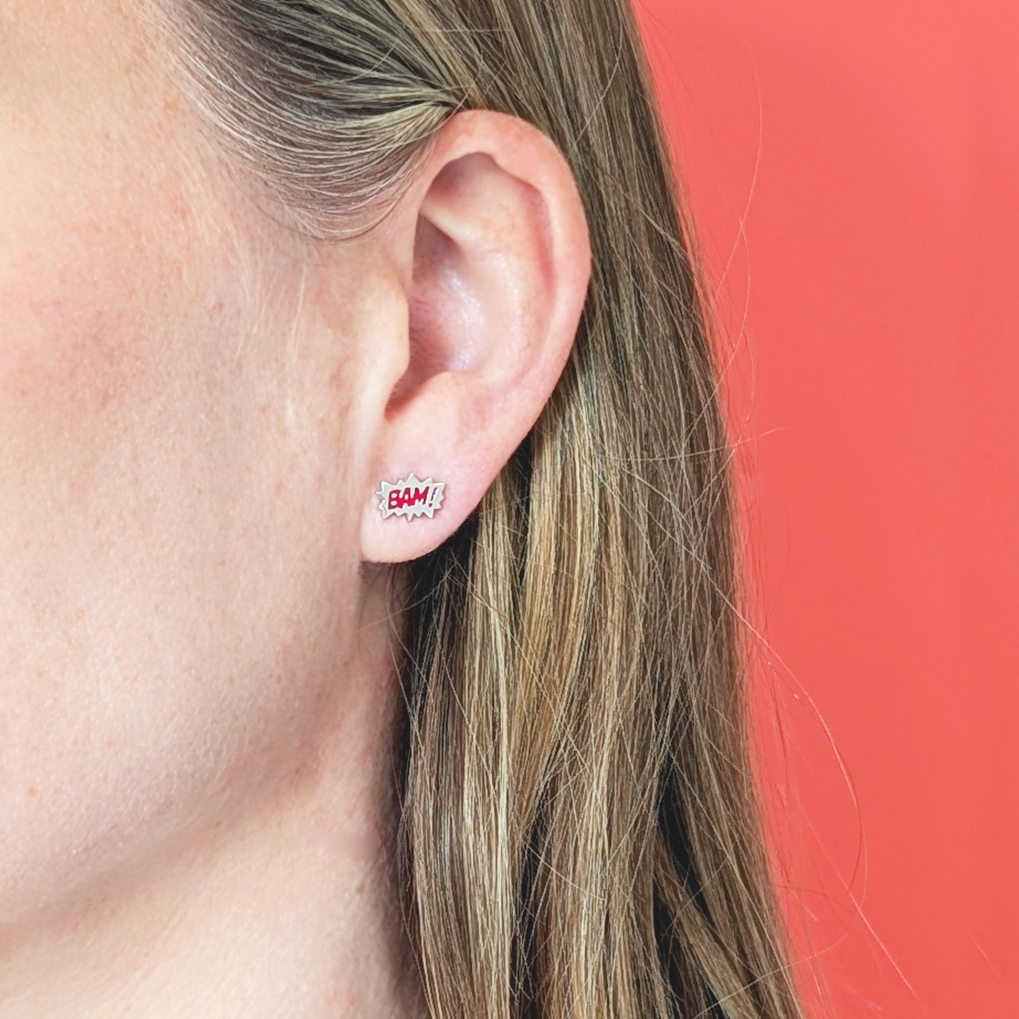 Close-up of an ear wearing a silver comic burst earring with red BAM! on a coral background