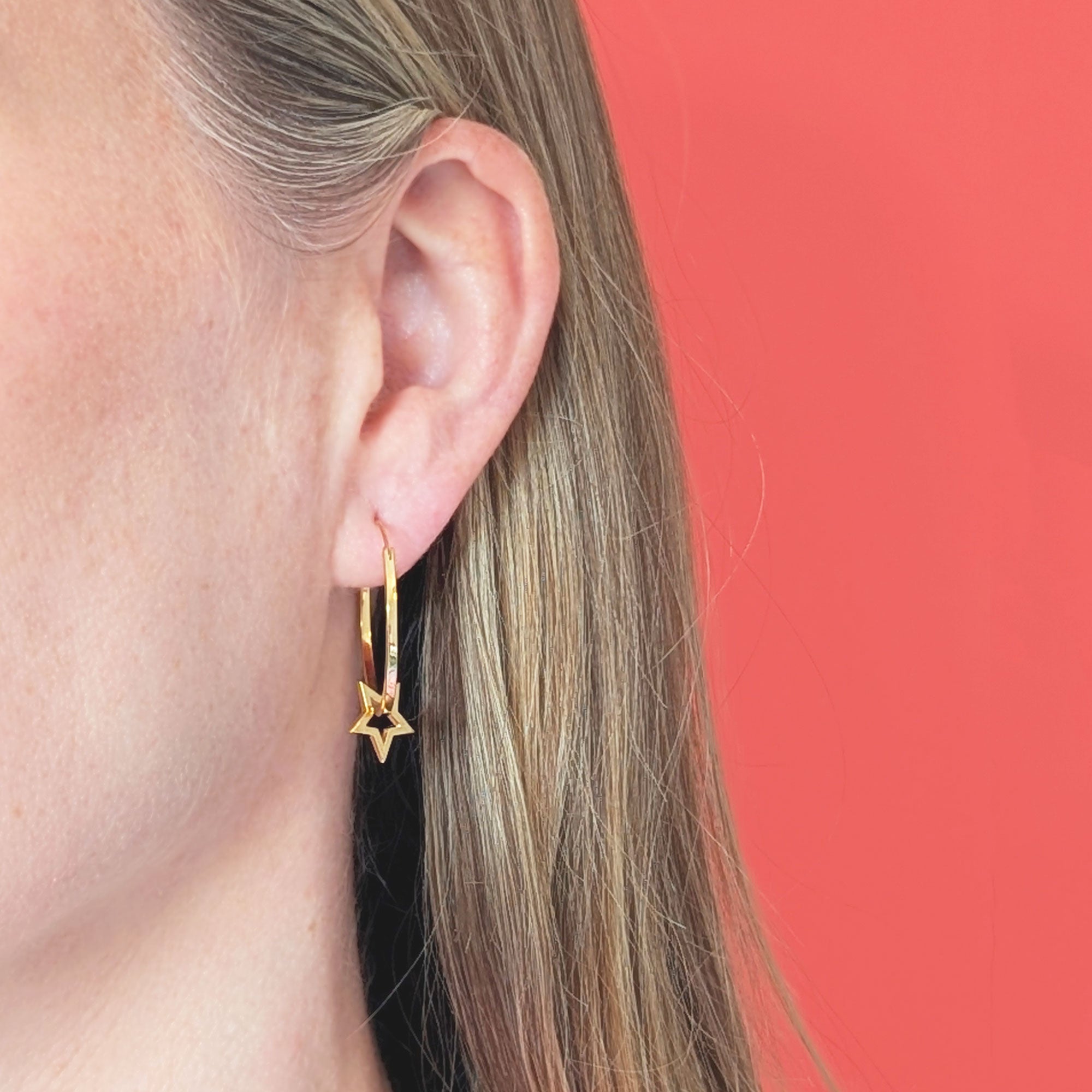 Gold hoop earrings with star-shaped charm threaded on worn by a person with a coral background