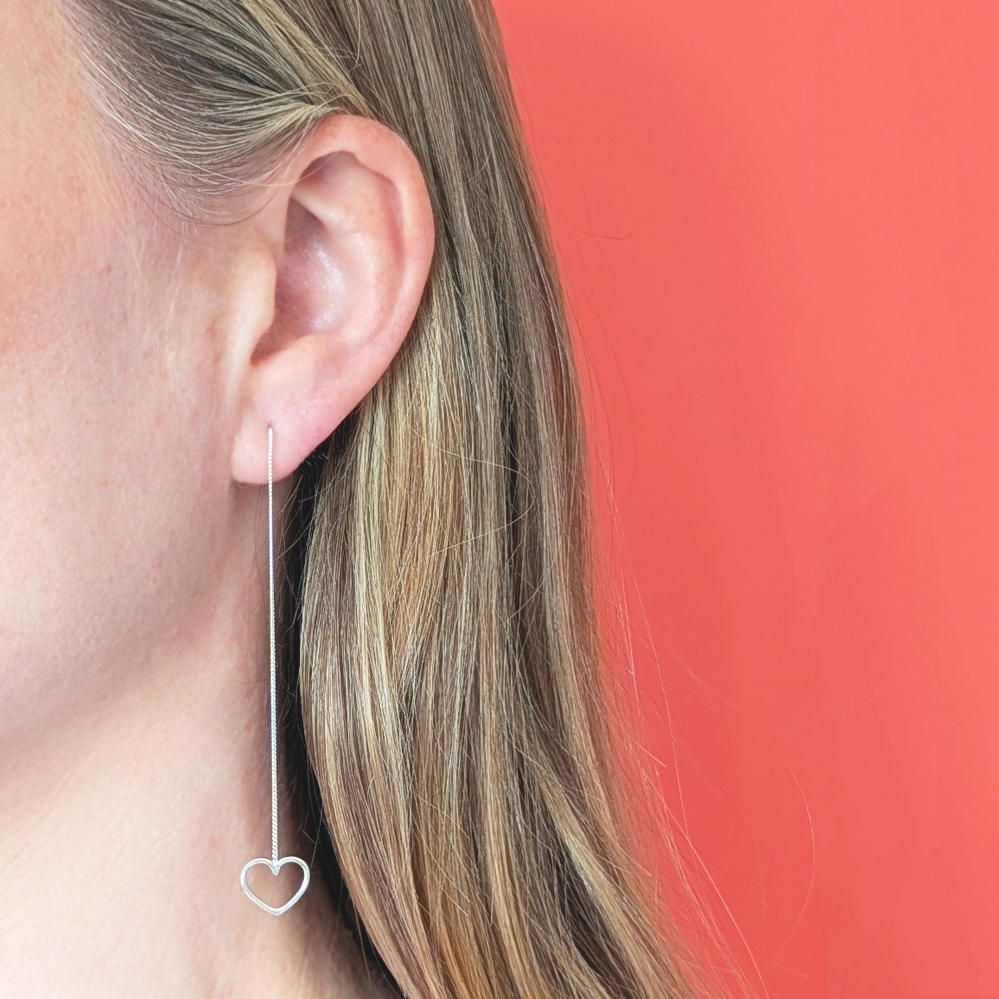 Silver heart-shaped chain threader earring worn by a person with a coral  background