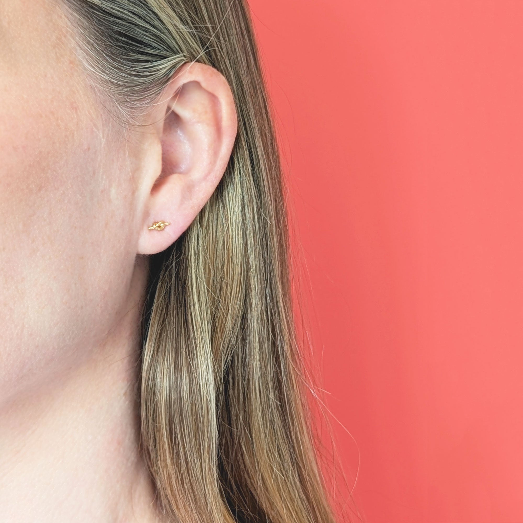 Close-up of an ear wearing a gold knot earring against a coral background