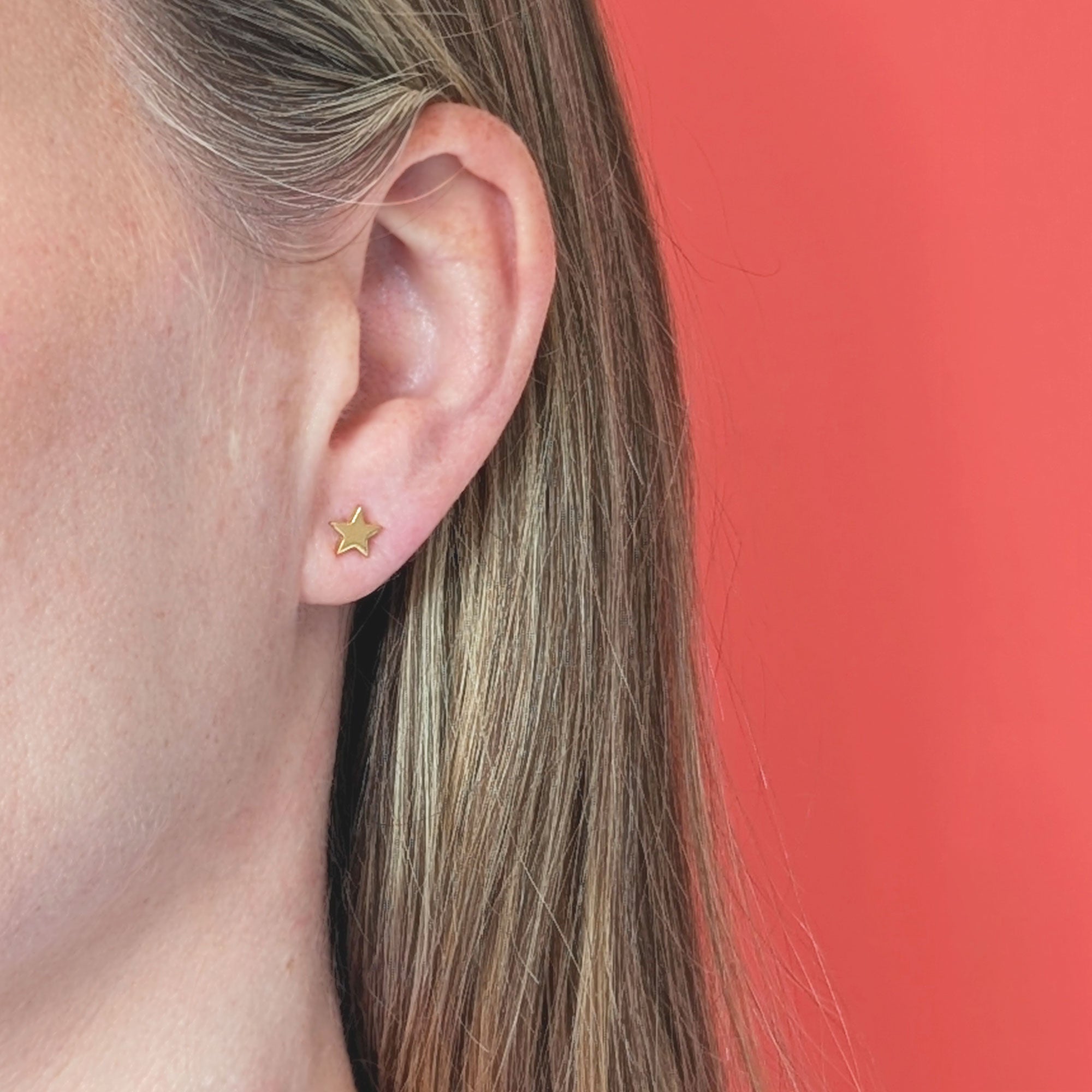 Gold star stud earring worn by a person with a coral background